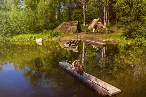 En person paddlar en stockbåt över en spegelblank sjö, omgiven av grönskande skog och historiska hyddor.