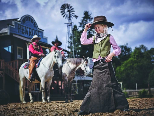 En flicka i westernkläder hälsar glatt med cowboyhatt på huvudet, medan cowboys rider i bakgrunden.