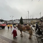 Häst och vagn med kusk i gammaldags kläder under Falköpings julparad. På torget syns en stor julgran, marknadsstånd och en traktor med en dekorerad vagn som följer paradtåget. Människor i julkläder och varma ytterkläder deltar i festligheterna.