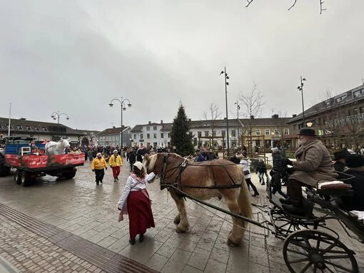 Häst och vagn med kusk i gammaldags kläder under Falköpings julparad. På torget syns en stor julgran, marknadsstånd och en traktor med en dekorerad vagn som följer paradtåget. Människor i julkläder och varma ytterkläder deltar i festligheterna.