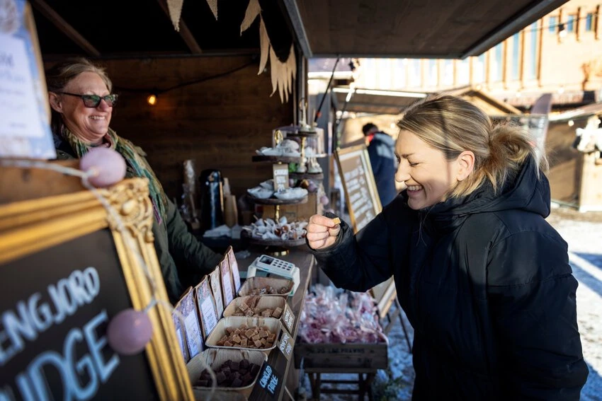 En glad besökare smakar på fudge vid ett marknadsstånd under torghandeln på Hertig Johans torg i Skövde. Försäljaren ler bakom disken där olika smaker av fudge är upplagda i korgar. Bakgrunden visar fler stånd och en solbelyst torgmiljö.